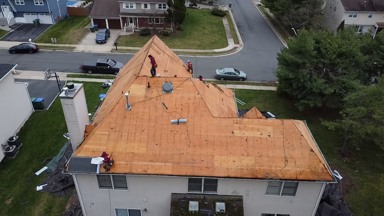 Aerial of a corner lot home with the entire roof stripped to bare plywood decking, crew members working on the open deck